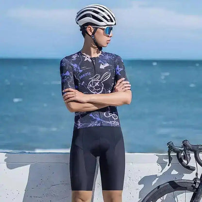 Mann i lilla Santic sykkeltrøye med grafisk mønster, sort sykkelshorts, hvit hjelm og sportsbriller ved havet.
