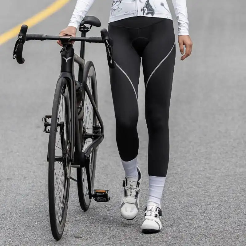 Kvinne i svart sykkeltights med hvite detaljer står ved siden av en landeveissykkel på asfalt.