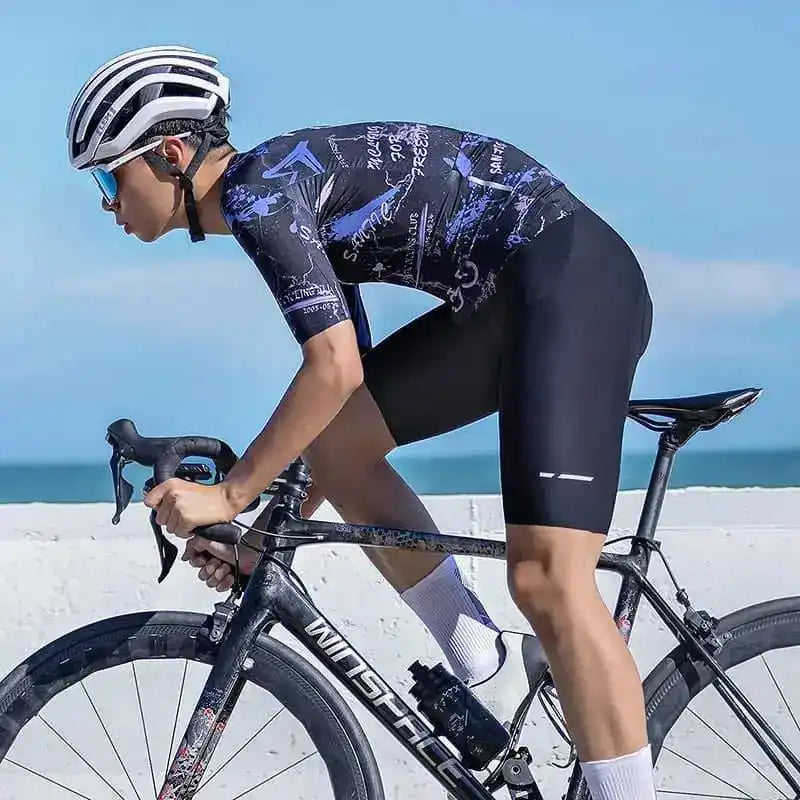 En mannlig syklist i mønstret sykkeltrøye og sorte bib-shorts på en Winspace landeveissykkel ved kysten.